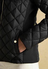 Čierna prešívaná bunda so zipsom vpredu, vyrobená z textúrovanej látky, s bočnými vreckami so zlatým kovaním a elastickými bočnými panelmi.