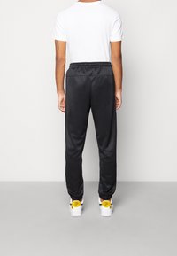 Man i svarta joggingsbyxor, vit T-shirt och vita sneakers med gula hälflikar som står vända mot en vit vägg på grått golv.