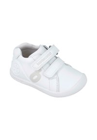 Zapatilla blanca de cuero con dos correas de velcro, puntera redondeada, suela texturizada y un logo circular en el lateral. Diseño minimalista.