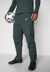 Tamnozeleni Nike hlače s bočnim džepovima na cipzaru, tkanina koja odbija vlagu i Chelsea FC logo na kuku, u kombinaciji s crnim kopačkama.