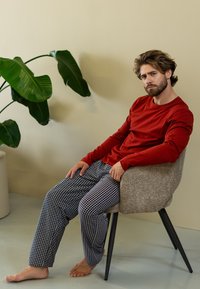 Rode shirt met lange mouwen gecombineerd met gestreepte marineblauwe broek, zittend in een gestructureerde grijze stoel, tegen een neutrale achtergrond met een plant.