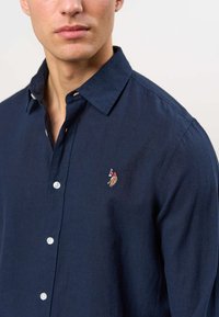 Muž nosi tmavomodrú košeľu na gombíky s malým vyšívaným logom hráča póla na ľavej strane hrude.