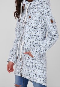 Lekka kurtka z kapturem o białej bazie w niebieskie i granatowe kropki. Posiada zamek błyskawiczny z przodu, sznurek oraz boczne kieszenie.