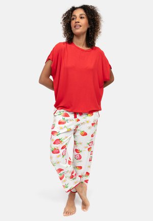 Donna in piedi a piedi nudi che indossa un top rosso largo e pantaloni bianchi con una stampa vivace di fragole rosse e fiori.