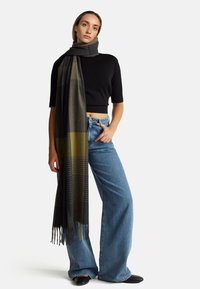 Kobieta w czarnym topie krótki, szerokich niebieskich dżinsach, czarnych butach oraz długim, ciemnoszarym i oliwkowym szalu owiniętym wokół szyi.