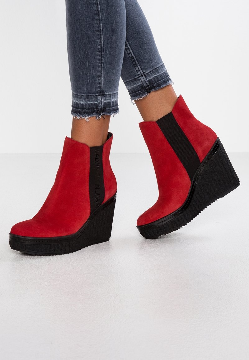 Czerwone zamszowe botki na koturnie z czarnym elastycznym panelem po boku, teksturowaną gumową podeszwą i gładkim, zaokrąglonym noskiem.