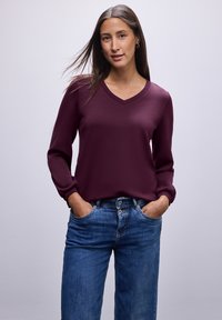 Burgundské triko s dlouhým rukávem a výstřihem do V, s nabíranými manžetami, spárované s modrými džínami. Hladká textura a uvolněný střih.