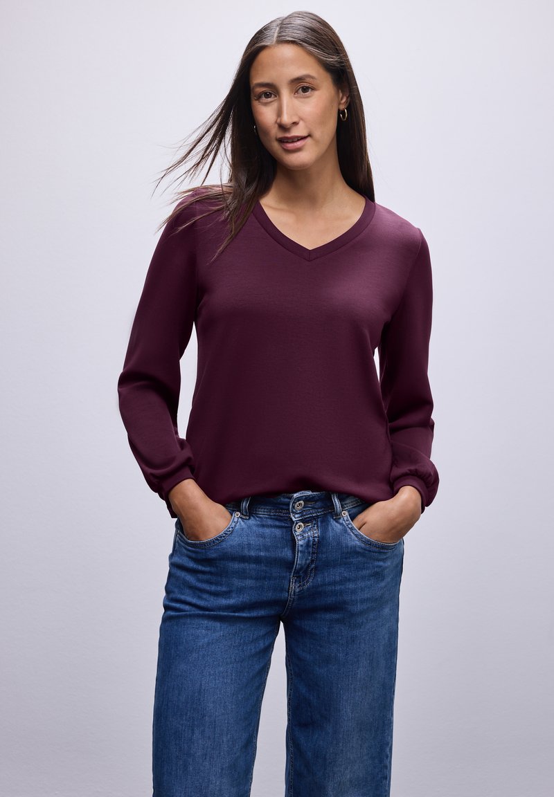 Burgunderfarbene Langarmbluse mit V-Ausschnitt und gerafften Manschetten, kombiniert mit blauen Jeans. Glatte Textur und lockere Passform.