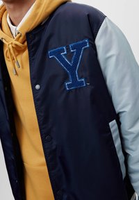 Marinblå och ljusgrå jacka med en framträdande blå broderad "Y"-logotyp, tryckknappar, bärs över en gul hooded sweatshirt.