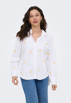 Jeune femme aux longs cheveux foncés portant une chemise blanche à boutons avec des motifs de soleil jaunes et un jean bleu, marchant vers l'avant sur un fond uni.