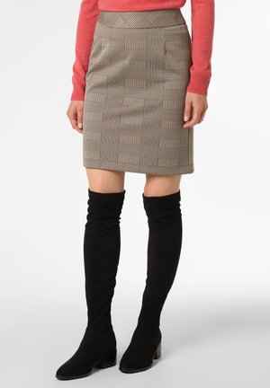 Frau trägt einen beige karierten knielangen Rock, ein korallenfarbenes Langarmshirt und schwarze Overknee-Stiefel aus Wildleder und steht vor weißem Hintergrund.