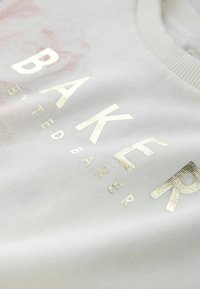 Sweat-shirt en coton blanc arborant l'inscription "BAKER" en doré métallisé, avec une finition texturée et un motif floral subtil en arrière-plan.