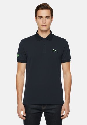 Modello maschile che indossa una polo nera con un triangolo verde e il logo "ΔX" sul petto, con testo sulla manica sinistra, in piedi su sfondo bianco.