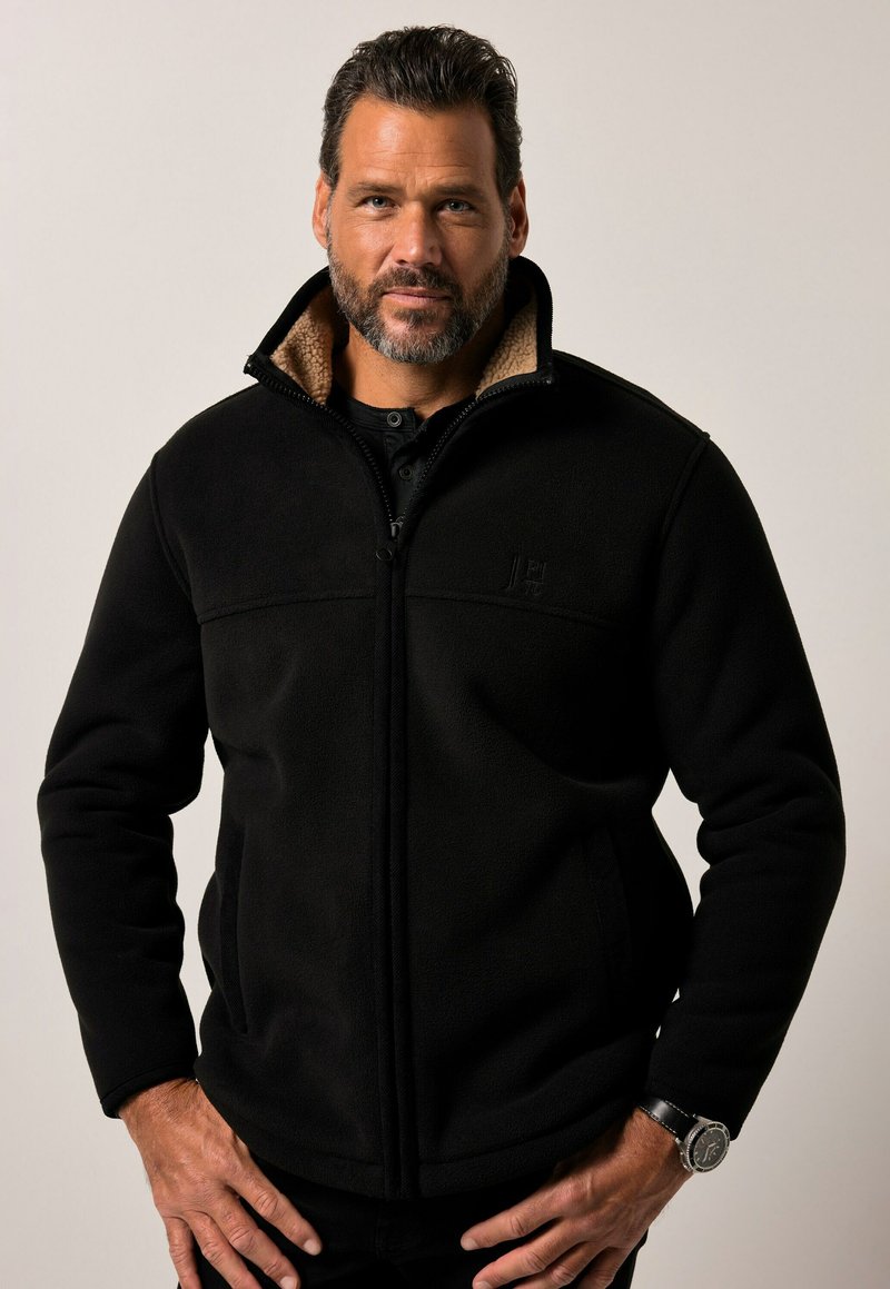 Chaqueta de forro polar negra con cierre de cremallera, cuello alto y forro marrón claro. Presenta bolsillos laterales y un logo sutil en el pecho.