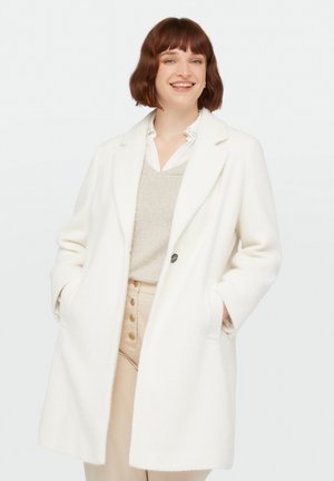 Cappotto bianco lungo in lana con rever, chiusura a un bottone e tasche laterali. Indossato sopra un maglione beige e pantaloni chiari.