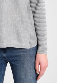 Grauer Pullover mit gerippten Ärmeln und glattem Körper, kombiniert mit blauen Jeans. Die Materialien wirken weich und das Design ist lässig.