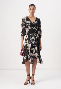 Robe noire enveloppante avec un motif floral en crème et beige. Tissu léger et transparent, décolleté rond et manches bouffantes. Accentuée avec un sac à main bordeaux.