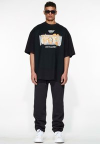 Camiseta negra de gran tamaño con estampado gráfico que dice "WASTED" y motivos de dólares. Combinada con pantalones joggers gris oscuro y zapatillas blancas.