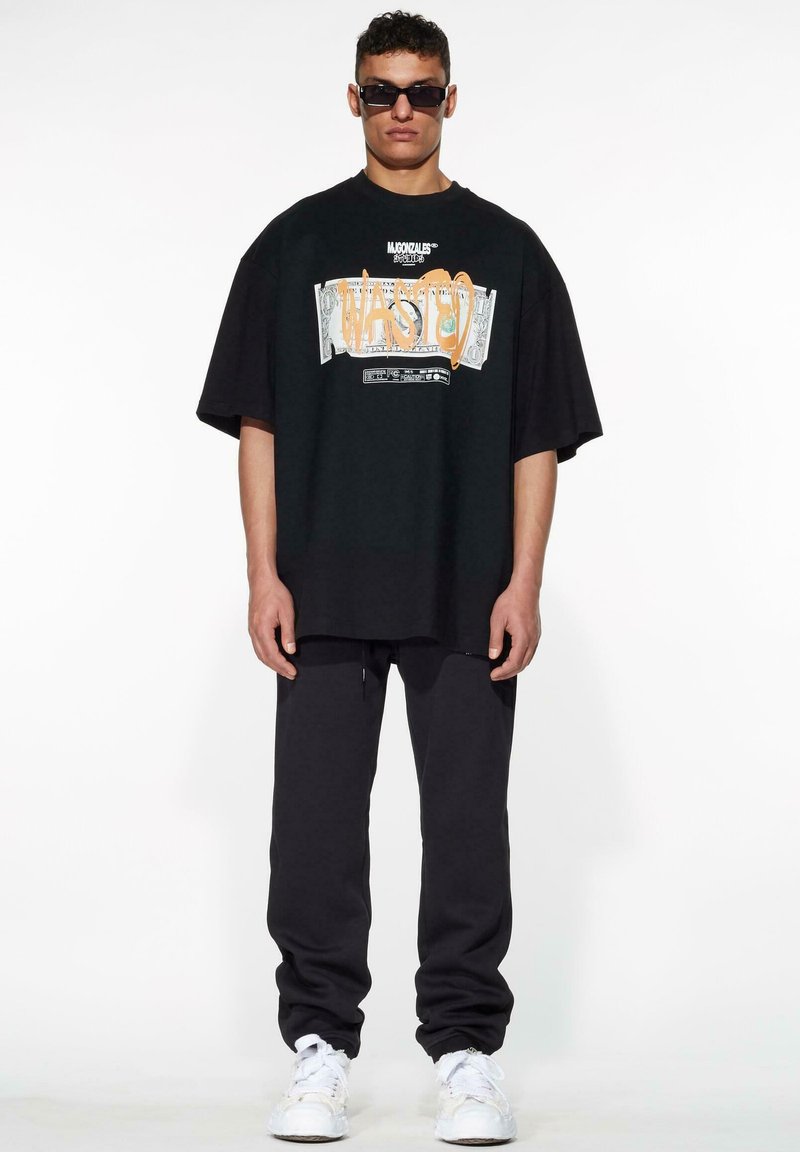 Camiseta negra de gran tamaño con estampado gráfico que dice "WASTED" y motivos de dólares. Combinada con pantalones joggers gris oscuro y zapatillas blancas.