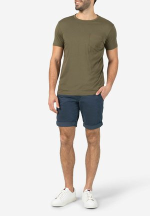 Olivegrünes T-Shirt mit Brusttasche, kombiniert mit navyblauen Shorts und weißen Sneakers. Einfaches Design und lässige Passform.