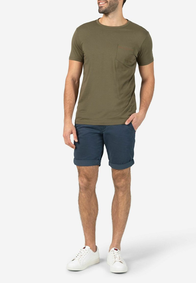 Olivegrünes T-Shirt mit Brusttasche, kombiniert mit navyblauen Shorts und weißen Sneakers. Einfaches Design und lässige Passform.