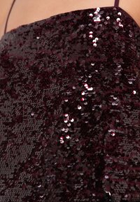 Tissu en sequins bordeaux à texture brillante, comportant de nombreux sequins circulaires qui reflètent la lumière, créant un effet scintillant.