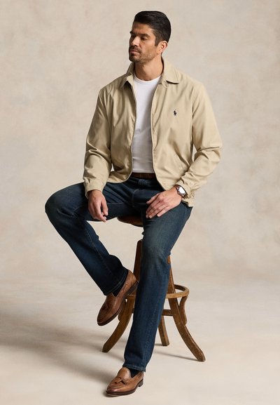 Mand sidder på en træskammel iført beige jakke, hvid t-shirt, mørke jeans, brune loafers og armbåndsur mod en neutral baggrund.