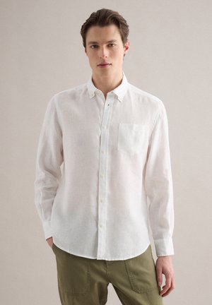 Junger Mann trägt ein weißes, langärmliges Hemd mit Knöpfen und olivgrüne Hose, steht mit einer Hand in der Tasche vor einem einfarbigen Hintergrund.