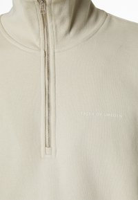 Sudadera beige con cremallera que presenta un alto cuello, textura acanalada y un pequeño logo "TIGER OF SWEDEN" en blanco en la parte izquierda del pecho.