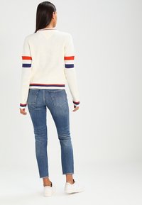 Suéter blanco de canalé con rayas rojas, azules marino y naranjas en las mangas y el dobladillo, combinado con jeans ajustados azules y zapatillas blancas.