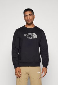 Sweatshirt negru cu decolteu round, cu un text mare de logo gri "THE NORTH FACE" și un accent curbat; afișat cu pantaloni khaki.