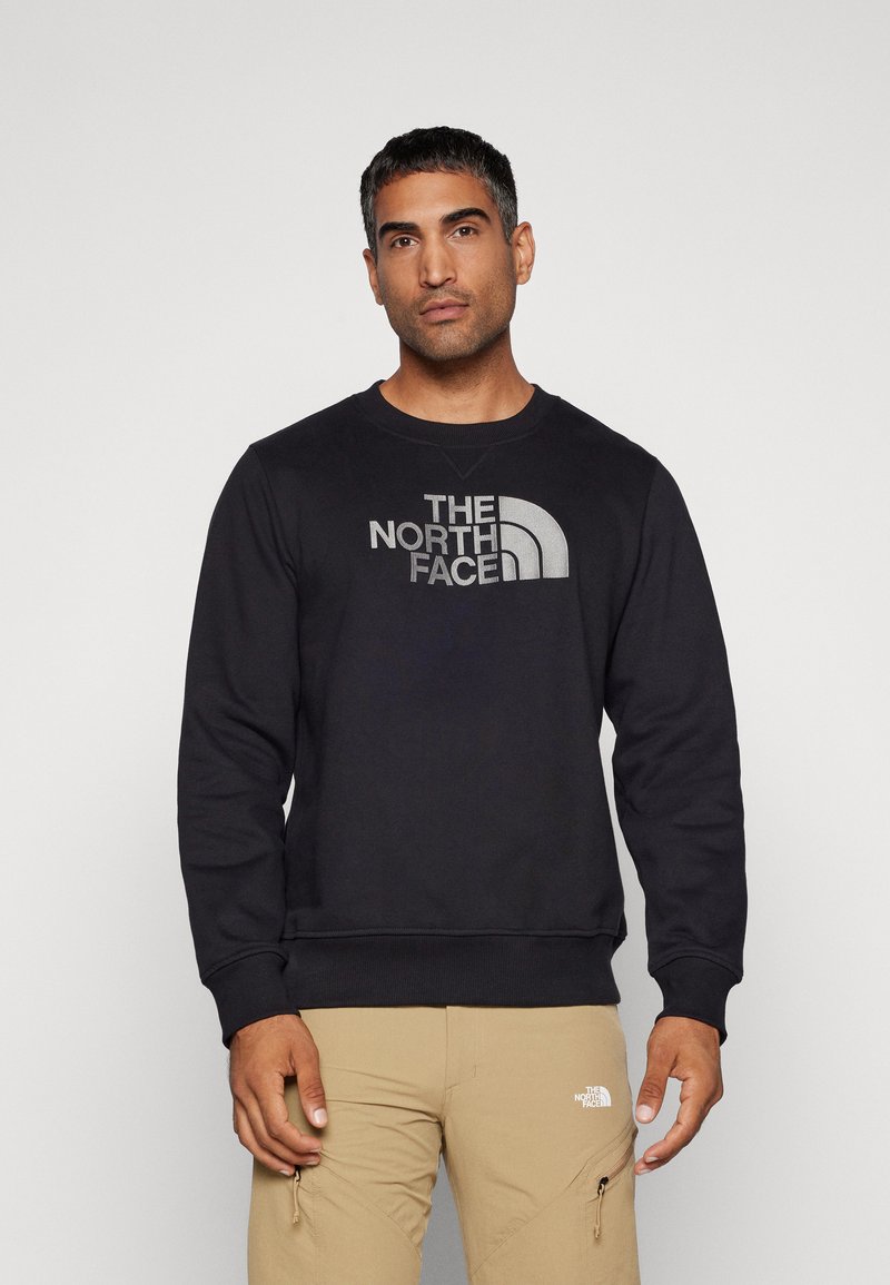 Sweatshirt negru cu decolteu round, cu un text mare de logo gri "THE NORTH FACE" și un accent curbat; afișat cu pantaloni khaki.