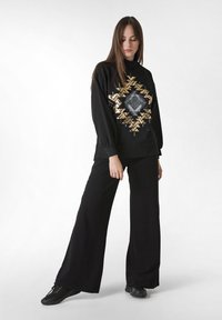 Svart oversized sweatshirt med guld geometriskt mönster, parat med vida svarta byxor och svarta skor. Slät tyg, avslappnad passform.