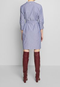 Modrá a bílá pruhovaná košilová šaty s dlouhými rukávy, zúženým pasem a tkaničkou. Outfit dotvářejí vínové kozačky až po kolena.