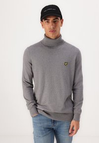 Sivi turtleneck pulover izrađen od mekanog materijala, s uskim krojem, rebrastim manžetama i malim žutim logotipom na lijevoj strani prsnog koša.