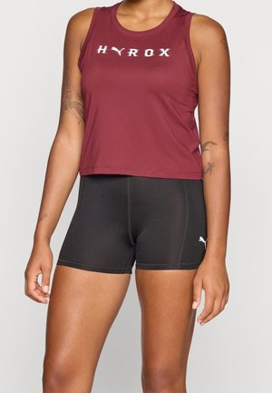 Vrouw draagt een mouwloos bordeauxrood HYROX crop top en zwarte Puma sportshorts, toont tatoeages op beide armen.