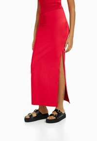 Czerwona, maxi sukienka do kolan z bocznym rozcięciem, wykonana z gładkiego materiału, zestawiona z masywnymi czarnymi sandałami z podwójnym zapięciem.