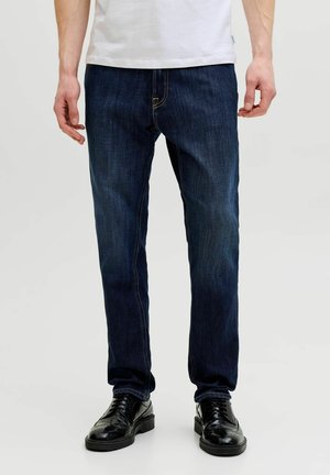 Mann trägt dunkelblaue Jeans mit geradem Schnitt und schwarze Lederschuhe, steht vor einem schlichten hellen Hintergrund.
