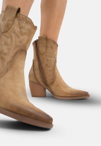 Botas de vaquero de cuero marrón claro con puntas afiladas, cremalleras laterales y patrones florales en relieve, con un tacón de madera apilada.