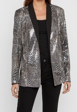 Blazer dengan pola python silver dan hitam dengan rona mengkilap, lapel hitam, dan desain terstruktur, dipadukan dengan atasan dan celana hitam.