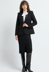 Černý upravený blazer s saténovými klopami, kombinovaný s černou sukní po kolena. Bílý motýlek a lesklé černé boty do kolen.