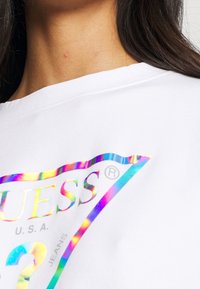 T-shirt en coton blanc présentant un grand imprimé logo irisé avec des accents bleus et arc-en-ciel à l'avant. L'encolure est large et décontractée.