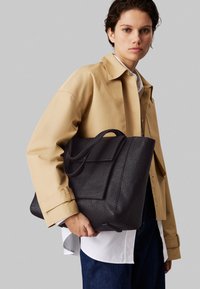 Black textured tote bag with dual handles and a front pocket. Worn with a beige jacket over a white shirt, set against a gray background.