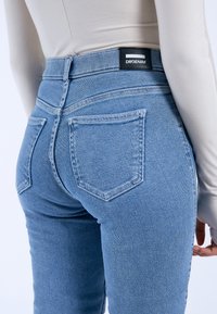 Blue denim jeans with a slim fit, featuring five pockets, a high waist, and a black label at the back. Textured fabric with minimal fading.