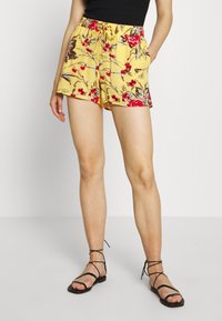 Pantalones cortos amarillos con un cinturón elástico, que presentan patrones de flores rojas y marrones. Combinados con sandalias negras de tiras.