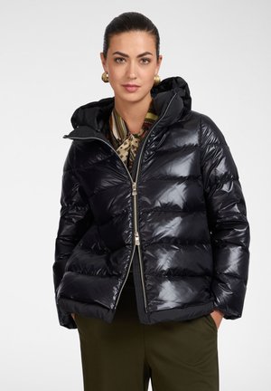 Frau mit dunklem Haar im glatten Dutt, die goldene Ohrringe, einen gemusterten Schal, eine glänzende schwarze Daunenjacke und grüne Hose trägt und mit den Händen in den Taschen steht.