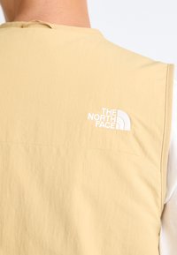 Light beige sleeveless vest in a soft fabric. Features a white embroidered logo on the upper left side. Simple design with minimal seams.