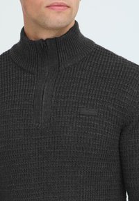 Mørkegrå zip-up sweater med et struktureret strikkemønster, ribbet krave og et lille sort logo på fronten.