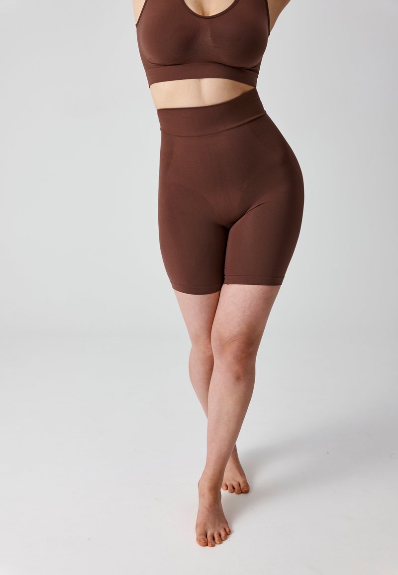 Woman wearing matching brown seamless sports bra and high-waisted biker shorts, standing barefoot on a white background.