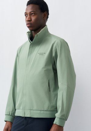 Άνδρας που φοράει ανοιχτό πράσινο μπουφάν με φερμουάρ, με όρθιο γιακά και τσέπες με φερμουάρ στα πλευρά, στέκεται μπροστά σε απλό λευκό φόντο.